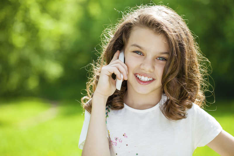 美丽的小女孩在夏日公园的背景下用手机打电话