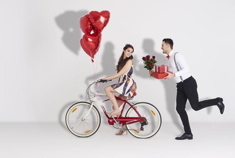 骑着自行车的美女身后有个拿着花和礼物的男人
