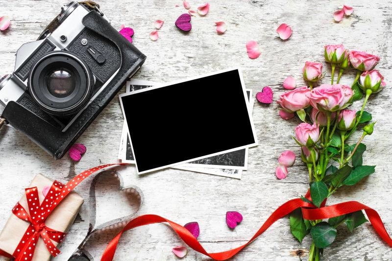 白色木桌上玫瑰花旁的礼物盒黑色老式照相机和空白相片