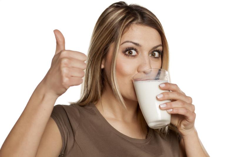 白色背景下竖起大拇指的快乐的年轻女子从杯子里喝牛奶