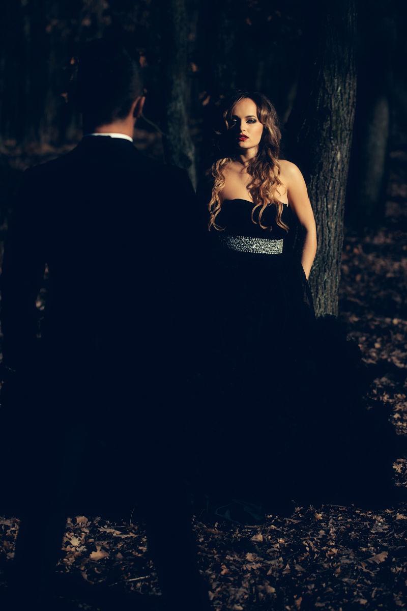森林中身着黑色西装的英俊男子和身穿优雅的晚礼服的性感女郎