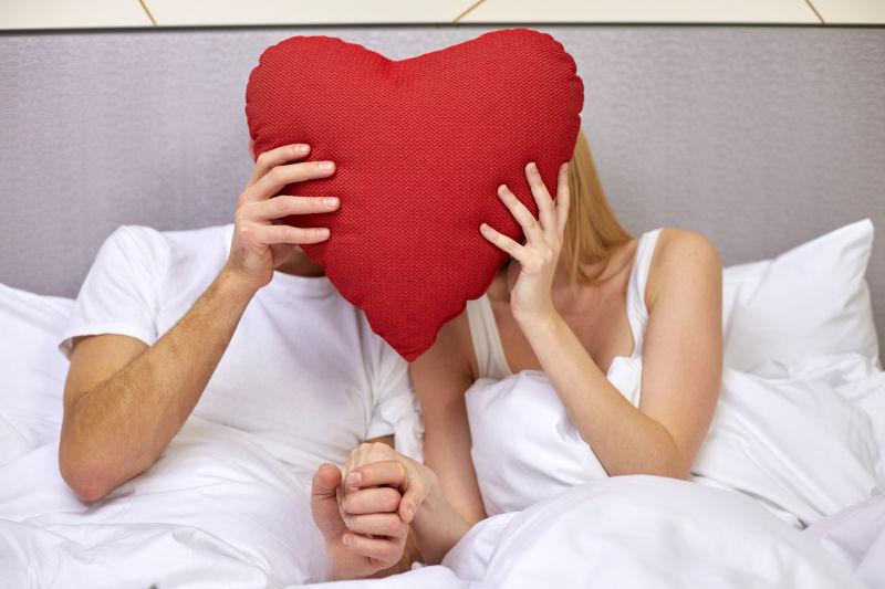 幸福的情侣在床上隐藏在红心枕头后面的旅馆或家中的面孔