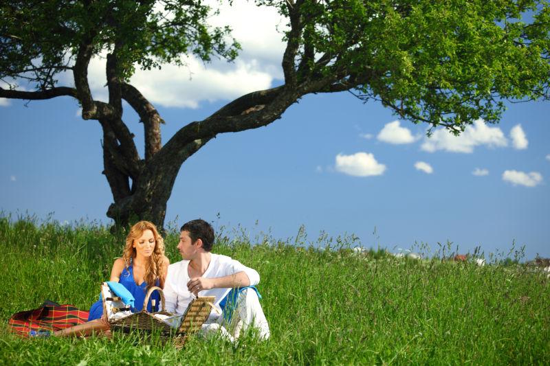 蓝天白云下大树下坐在草地上吃东西的情侣
