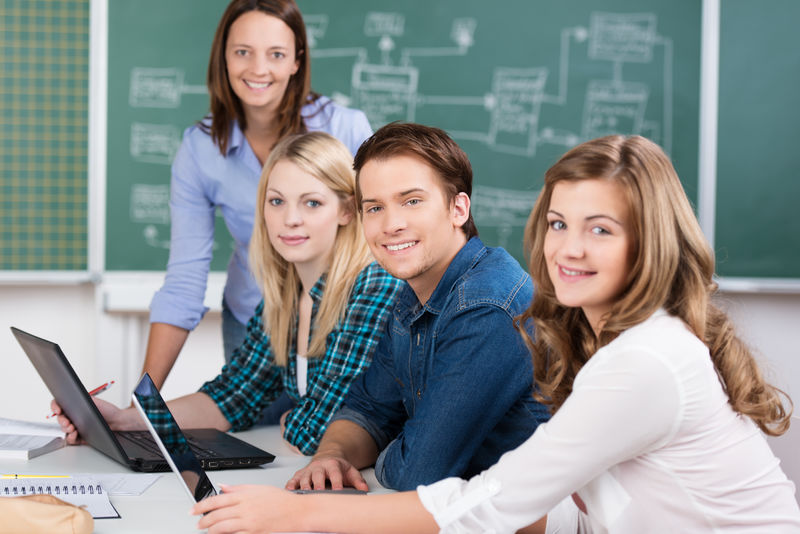 在大学教室里-一群有魅力的青少年学生坐在桌子旁-笔记本电脑放在桌上-一位女教师站在教室的尽头