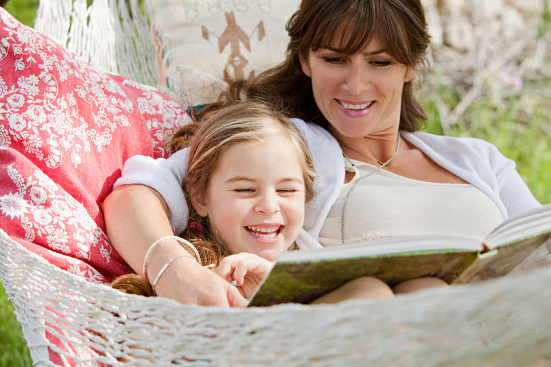 一位美丽的年轻母亲和她五岁的女儿躺在吊床上-在阳光明媚的夏日里顽皮地读着一本儿童故事书-户外家庭生活