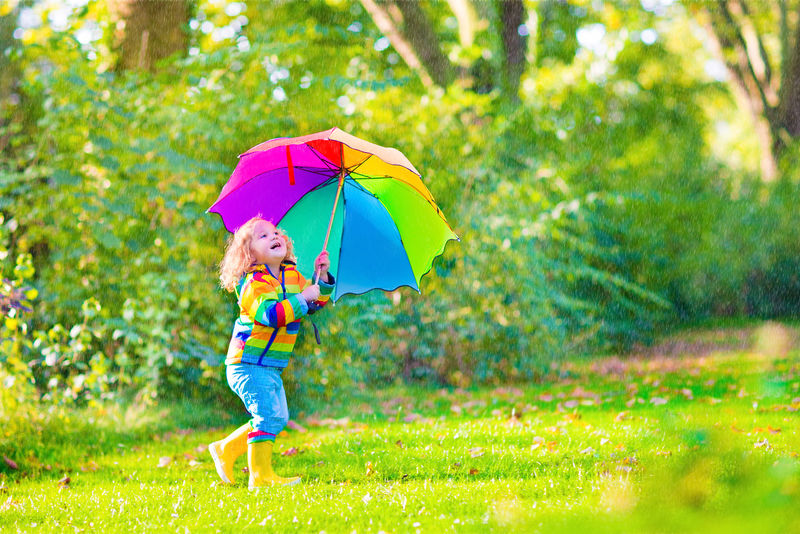 一个可爱的小女孩-穿着黄色防水外套和靴子-在温暖的秋日或夏天-在雨中和阳光下-手持五颜六色的雨伞在花园里玩耍