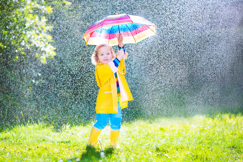 一个可爱的小女孩-穿着黄色防水外套和靴子-在温暖的秋日或夏天-在雨中和阳光下-手持五颜六色的雨伞在花园里玩耍