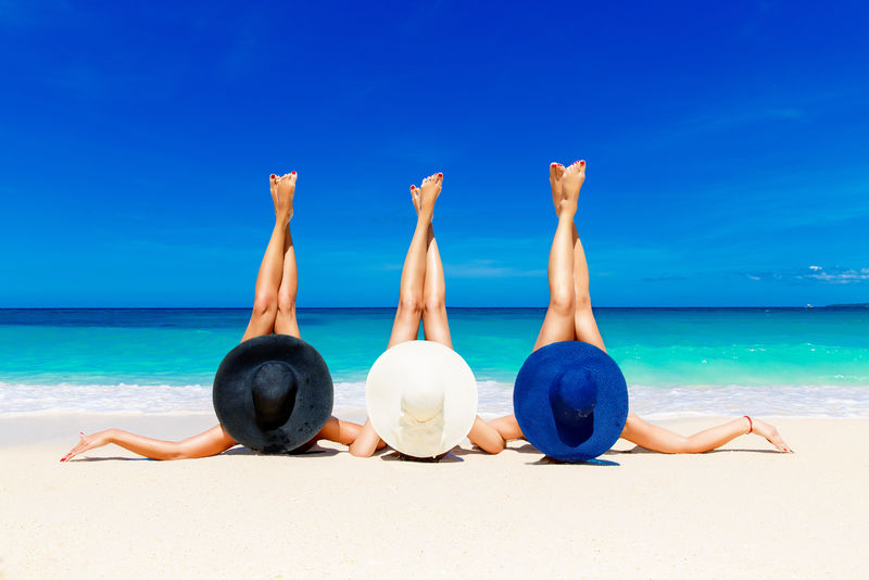 三个戴草帽的年轻女子躺在热带海滩上-伸开纤细的腿-背景是蓝海-暑假概念