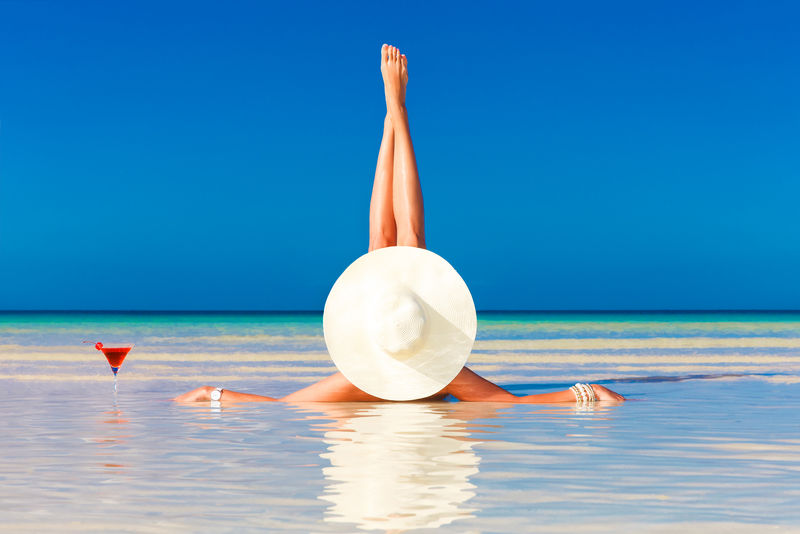 戴草帽的年轻女子躺在热带海滩上享受鸡尾酒