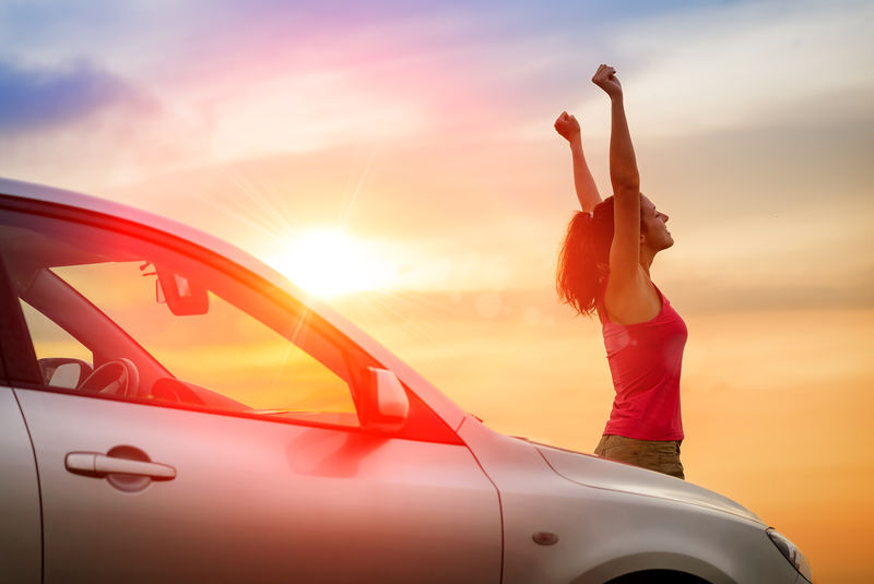 坐在车旁的女司机举起双臂-感受着朝着日落方向自由驾驶的感觉-妇女和车辆在美丽的阳光背景下