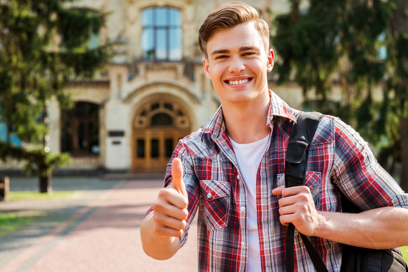 竖起大拇指争取成功！一位英俊的男学生，站在户外，背景是大学大楼，他抬起大拇指微笑着。