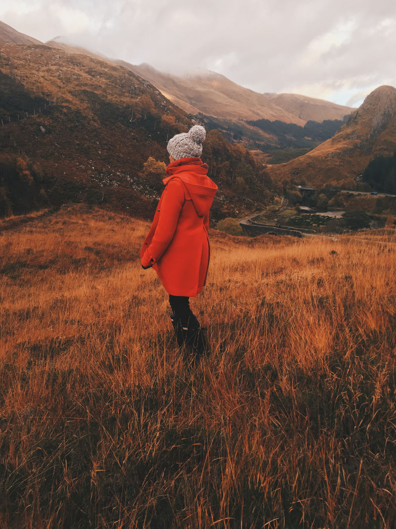 穿着橙色外套的女人站在山边的草地上