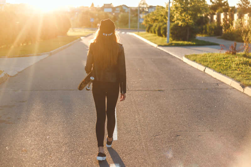 阳光下拿着滑板散步的年轻女孩背影
