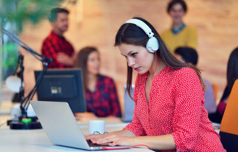 现代办公室背景下的带着耳机用笔记本工作的女人